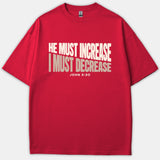A red Christian t-shirt featuring the bold text HE MUST INCREASE in white and I MUST DECREASE in beige, with the Bible verse reference JOHN 3:30 underneath.