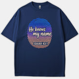 A navy blue Christian t-shirt featuring a colorful gradient fingerprint graphic with the phrase 'He knows my name' and scripture reference 'ISAIAH 43:1' in cream script.