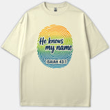 A beige Christian t-shirt featuring a colorful fingerprint design with the phrase He knows my name and scripture reference Isaiah 43:1.