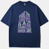 A navy blue Christian t-shirt featuring 3D tower-shaped typography displaying the Bible verse, 'The name of the Lord is a strong tower,' in green and purple lettering.