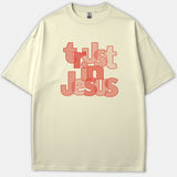 A beige t-shirt featuring overlapping typography that reads 'trust in Jesus' printed in shades of red and pink.