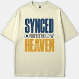 A beige Christian t-shirt featuring bold typography that reads 'SYNCED WITH HEAVEN' alongside media-themed icons in navy blue, gray, and gold.