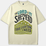 A beige Christian t-shirt featuring graphic typography reading 'The Lord Is My Shepherd' above an illustration of a shepherd with sheep grazing on a green hill.