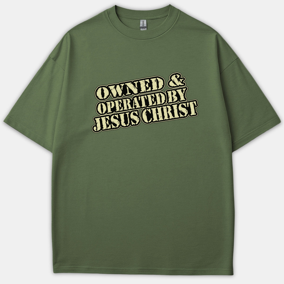 An olive green Christian t-shirt featuring the text OWNED & OPERATED BY JESUS CHRIST printed across the chest in a vintage beige font with black outlines.