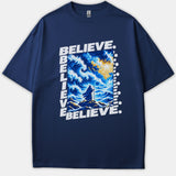 A navy blue t-shirt featuring a dramatic graphic of a robed figure before parting ocean waves under divine sunlight, framed by the word BELIEVE in bold white text.