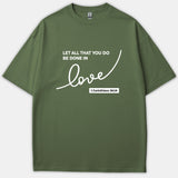 An olive green Christian t-shirt featuring the Bible verse 'Let all that you do be done in love' from 1 Corinthians 16:14 printed in white cursive and block lettering.