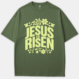 An olive green t-shirt featuring a light yellow floral and butterfly design centered around the bold text Jesus Is Risen.