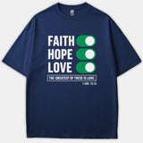 A navy blue Christian t-shirt featuring the words 'Faith, Hope, Love' next to green toggle switches, above the scripture phrase 'The Greatest of These is Love' and 1 Cor. 13:13.
