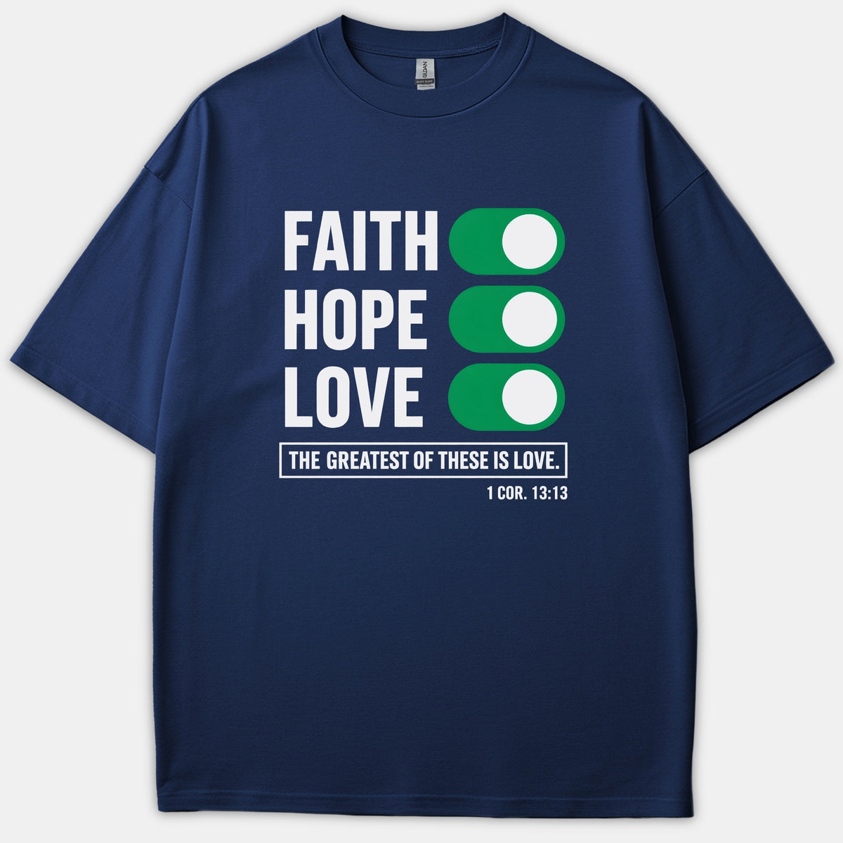 A navy blue Christian t-shirt featuring the words 'Faith, Hope, Love' next to green toggle switches, above the scripture phrase 'The Greatest of These is Love' and 1 Cor. 13:13.