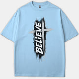 A light blue Christian t-shirt featuring a distressed black and gray cross graphic with the word BELIEVE printed vertically in bold white brush font.