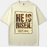 A beige Christian t-shirt featuring a scroll graphic with an empty tomb illustration and the scripture verse He Is Risen Just As He Said from Matthew 28:6.