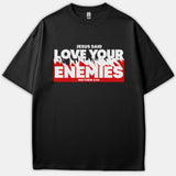 A black t-shirt featuring a bold graphic print that reads 'JESUS SAID LOVE YOUR ENEMIES MATTHEW 5:44' in white text over a distressed red background.