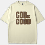 A beige Christian t-shirt featuring a graphic print that reads GOD IS GOOD in bold typography filled with a purple and gold wavy line pattern.