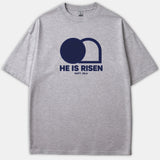 A gray Christian t-shirt featuring a navy graphic of an empty tomb with the text 'HE IS RISEN MATT. 28:6' printed on the chest.