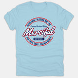 A light blue Christian t-shirt featuring a circular emblem graphic with the verse Matthew 5:7, 'Blessed Are The Merciful', printed in red, white, and navy blue.