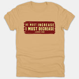 A beige Christian t-shirt featuring a red painted background graphic with the Bible verse 'He Must Increase I Must Decrease John 3:30' printed in light yellow text.