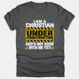 A dark gray t-shirt featuring a construction sign graphic with the text I Am A Christian Under Construction God's Not Done With Me Yet.