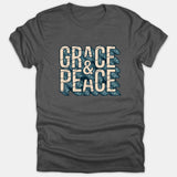 A dark gray Christian t-shirt featuring the phrase 'GRACE & PEACE' in bold beige block lettering against a blue wave graphic background.