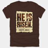 A dark brown Christian t-shirt featuring a graphic print reading 'He Is Risen, Just As He Said! Matt. 28:6' inside a parchment scroll with an empty tomb illustration.