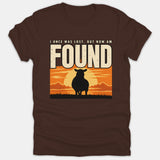 A brown Christian t-shirt featuring the silhouette of a sheep against a vibrant sunset with text that reads, 'I Once Was Lost, But Now Am Found'.