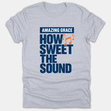 A heather gray Christian t-shirt featuring navy and orange typography that reads 'AMAZING GRACE HOW SWEET THE SOUND' accompanied by musical note graphics.