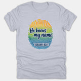 A gray Christian t-shirt featuring a colorful fingerprint graphic with the text 'He knows my name' and the scripture reference ISAIAH 43:1.
