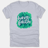 A light gray Christian T-shirt featuring the text 'have faith' in white cursive typography over a green tropical floral illustration.