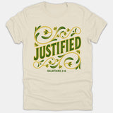 A beige Christian t-shirt featuring the word JUSTIFIED in green lettering surrounded by ornate gold and green floral flourishes above the scripture reference Galatians 2:16.