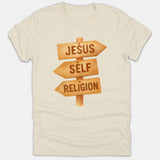 A beige Christian t-shirt featuring a graphic of a wooden signpost with directional arrows labeled Jesus, Self, and Religion.