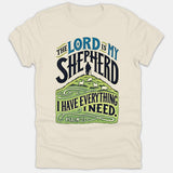A beige Christian t-shirt featuring a pastoral graphic illustration of a shepherd with sheep on a hill, surrounded by text from Psalm 23.
