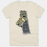 A beige t-shirt featuring a stylized Christian graphic with the phrase Simply Blessed in bold, distressed yellow and gray block lettering with paint splatters.