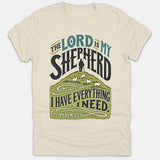 A beige Christian t-shirt featuring Psalm 23 scripture artwork that reads The Lord Is My Shepherd above an illustration of a shepherd leading sheep on a green hill.