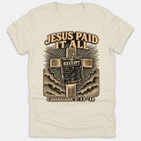 A beige Christian T-shirt featuring a graphic of a wooden cross with a receipt nailed to it that reads Paid in Full, surrounded by the words Jesus Paid It All and Colossians 2:13-14.