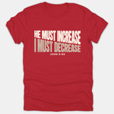 A red Christian t-shirt featuring the Bible verse 'He Must Increase I Must Decrease' from John 3:30 printed in bold white and beige lettering.
