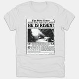 A white Christian t-shirt featuring a vintage newspaper graphic titled The Bible Times with the headline He Is Risen and an illustration of Jesus' empty tomb.