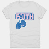 A white Christian t-shirt featuring hanging blue boxing gloves beneath pink and blue text that reads 'Fight the Good Fight of Faith 1 Timothy 6:12'.