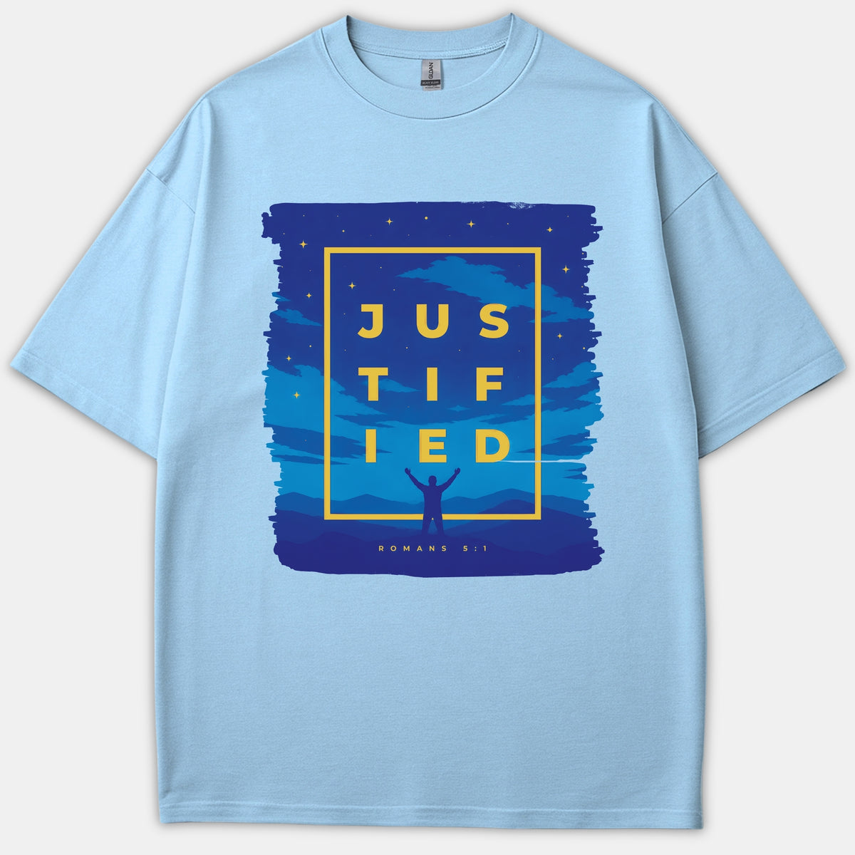 A light blue T-shirt with a navy graphic featuring stars, a silhouette raising hands, and yellow text reading JUSTIFIED along with the reference Romans 5:1.