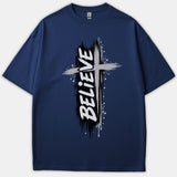 A navy blue Christian t-shirt featuring a distressed brushstroke cross graphic with the word BELIEVE printed vertically in bold white lettering.