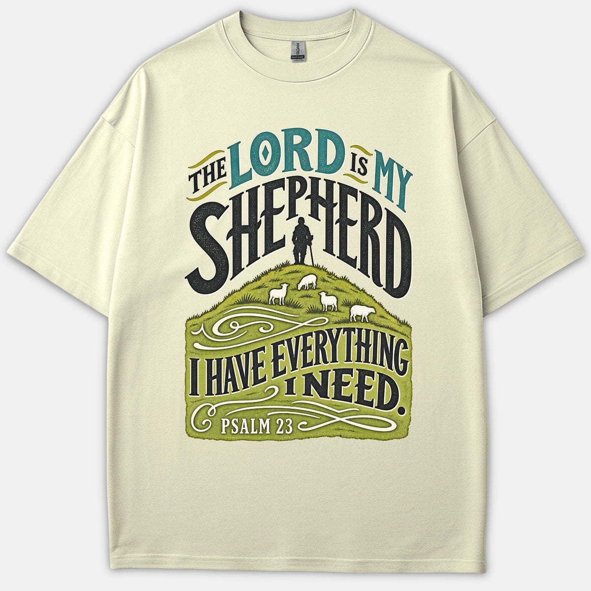 A beige Christian t-shirt featuring graphic typography reading 'The Lord Is My Shepherd' above an illustration of a shepherd with sheep grazing on a green hill.