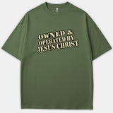 An olive green Christian t-shirt featuring the text OWNED & OPERATED BY JESUS CHRIST printed across the chest in a vintage beige font with black outlines.