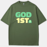 An olive green Christian t-shirt featuring the phrase GOD 1ST in bold mint green and light yellow lettering with a small cross graphic.