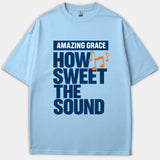 A light blue Christian T-shirt displaying the bold phrase Amazing Grace How Sweet The Sound in navy blue with vibrant orange musical notes.