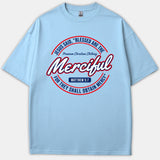 A light blue Christian T-shirt featuring a circular graphic with the Matthew 5:7 scripture quote, Blessed Are The Merciful, stylized in red, navy, and white lettering.