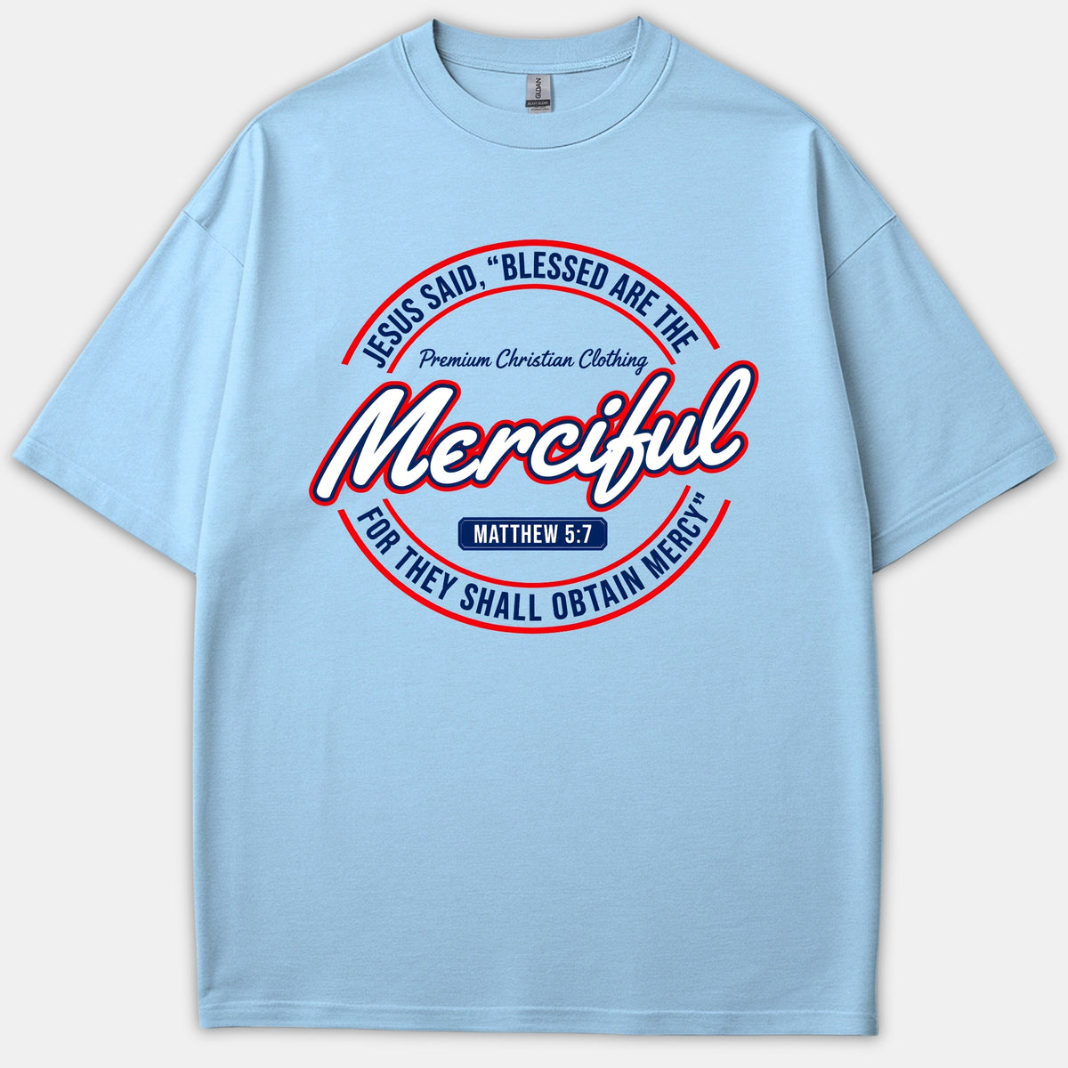 A light blue Christian T-shirt featuring a circular graphic with the Matthew 5:7 scripture quote, Blessed Are The Merciful, stylized in red, navy, and white lettering.