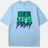 A light blue t-shirt featuring the phrase REAL MEN PRAY in a bold green and navy graphic paint-splatter typography.