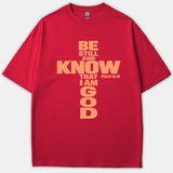 A red Christian t-shirt featuring the scripture phrase Be Still And Know That I Am God arranged in the shape of a cross with Psalm 46:10.