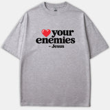 A heather gray t-shirt featuring a red heart icon followed by bold black text that reads 'your enemies - Jesus'.