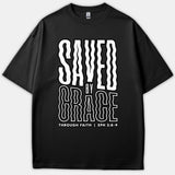 A black t-shirt featuring a wavy, retro-style graphic text that reads SAVED BY GRACE THROUGH FAITH with the scripture reference EPH 2:8-9.