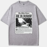A heather gray t-shirt featuring a vintage newspaper front-page design titled 'The Bible Times' with the main headline 'HE IS RISEN!' and an illustration of Jesus' empty tomb.