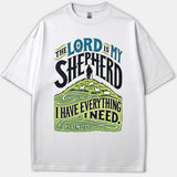A white Christian T-shirt featuring decorative typography reading 'The Lord Is My Shepherd' above an illustration of a shepherd and sheep on a green hill with Psalm 23.
